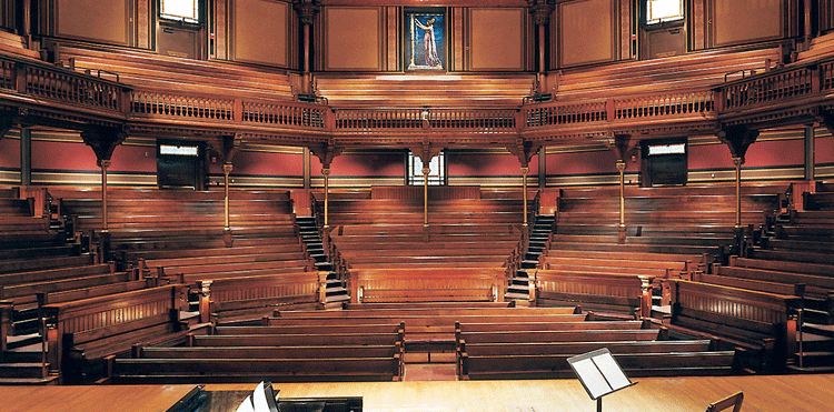 interior view of the semicircular wooden mezzanine and orchestra in Sanders Theatre, with a grand piano and music stand on stage